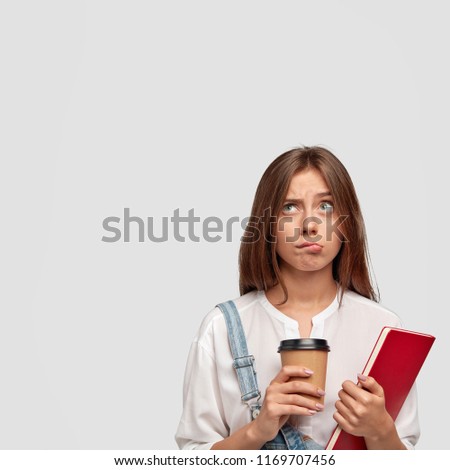 Indoor vertical shot of pleasant looking brunette young girl or student thinks on creation new project work, being involved in creative group, reads literature, has coffee break after lectures