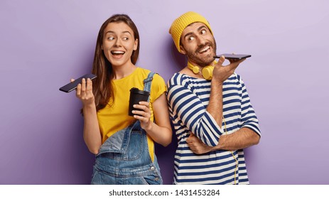 Indoor View Of Happy Woman And Man Keep Modern Cell Phones Near Mouthes, Make Voice Call, Enjoy Nice Conversation, Drink Hot Beverage From Disposable Cup, Stand Back To Each Other Over Purple Wall