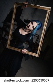 Indoors Portrait Of Lovely Goth Girl In Black Dress. Blue-haired Gothic Lady Holds A Picture Frame In Hands