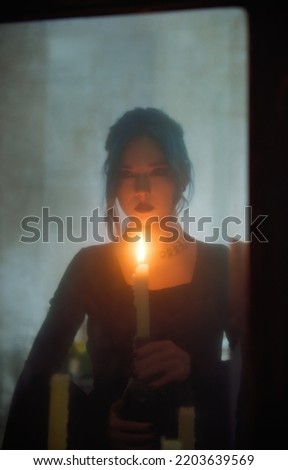 Indoors portrait of sad goth girl with candle. Blue-haired gothic lady looking into the old dirty mirror. Young witch. Vintage style