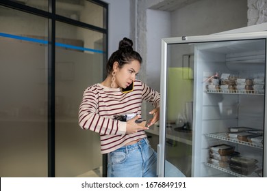 Indoors. Young Woman With Food Boxes Holding Fridge Door, Talking On Mobile Phone, Serious, Busy