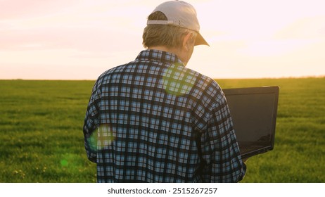 Industrial agrarian male farmer examining green wheat field plantation at sunset use laptop closeup. Modern technician agricultural scientist agronomist analyzing crop cultivation produce back view - Powered by Shutterstock