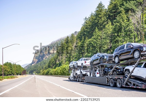 Industrial Big rig white classic car hauler semi\
truck tractor transporting cars on the modular hydraulic two level\
semi trailer driving on the one way highway road along the river in\
Columbia Gorge
