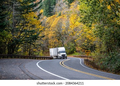 Industrial big rig white semi truck tractor with grille guard transporting commercial cargo in dry van semi trailer driving on the narrow winding road with autumn forest on the hill in Columbia Gorge