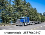 Industrial blue classic big rig semi truck tractor with extended cab for truck driver rest transporting empty flat bed semi trailer driving on wide highway road with trees on side in Columbia Gorge