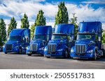 Industrial blue classic carrier big rig semi trucks tractors with extended cab and without semi trailers standing in row on industrial parking lot with green trees waiting for the next freights