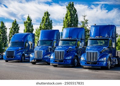 Industrial blue classic carrier big rig semi trucks tractors with extended cab and without semi trailers standing in row on industrial parking lot with green trees waiting for the next freights