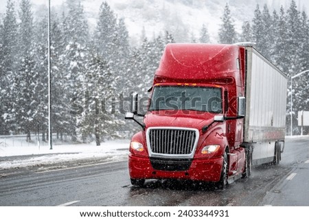 Industrial bright red big rig long hauler semi truck transporting cargo in dry van semi trailer cautiously driving on a dangerous winter highway during snowfall in Shasta Lake area in California