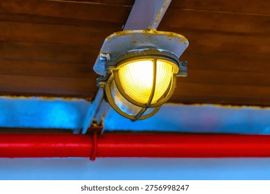 Industrial bulkhead ceiling light in selective focus with metal cage and warm yellow glow on wooden background.Vintage nautical ship lantern in industrial loft style near red metal pipe.