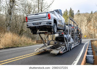 Industrial burgundy classic car hauler big rig semi truck with grille guard transporting cars on the two level hydraulic semi trailer running on the mountain winding road with hills on the sides