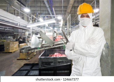 Industrial butcher posing with two filleting knives, wearing protective and hygienic clothing, such as a white suit, mask and a yellow hard hat, in front of a large animal processing plant - Powered by Shutterstock