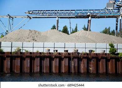 Industrial Canal River Scene Of Concrete Manufacturing Facility With Overhead Mechanical Conveyor Belt System Dumping Large Piles Of Sand And Gravel Beside Cement Wall And Old Rusted Pylon Sea Wall