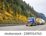 Industrial carrier big rig blue semi truck tractor with oversize load sign transporting part of oversized wind turbine pillar on semi trailer with trolley running on autumn road in Columbia Gorge 