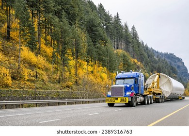Industrial carrier big rig blue semi truck tractor with oversize load sign transporting part of oversized wind turbine pillar on semi trailer with trolley running on autumn road in Columbia Gorge 