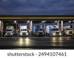 Industrial carriers big rig semi trucks with semi trailers fill the tanks with diesel fuel standing on the truck stop gas station with lights at night to be ready to continue the route by morning