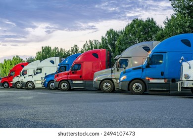 Industrial carriers big rig semi trucks with semi trailers standing for rest in row on the truck stop parking lot with turned on lights at twilight time to be ready to continue the route by morning