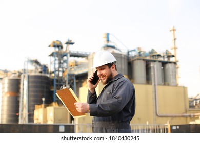 Industrial Caucasian Engineer Talking By VHF Walkie Talkie And Holding Papers Near Factory Outside. Male Wearing White Helmet And Blue Work Jumpsuit. Concept Modern Technology, Plant Profession And