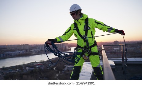 Industrial climber in a safety belt and a white helmet throws a rope while standing on the edge of a roof at sunset in slow motion