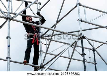 An industrial climber wearing a helmet and a protective belt is tied with a rope to a support at a high altitude. A specialist assembles a stage structure from a modular system of metal scaffolding.