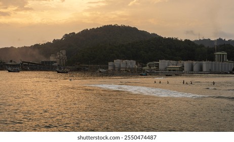 An industrial complex along a coastal shoreline at sunset, with silhouettes of people wading in the water and distant fishing boats, creating a contrast between nature and industry. - Powered by Shutterstock