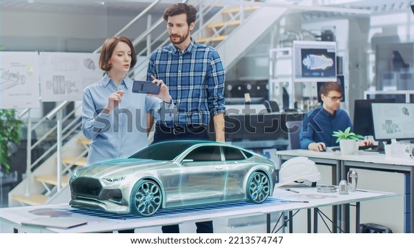 Industrial Design: Automotive Engineer and\
Designer Working Together on 3D Electric Car Design, Using\
Smartphone with Augmented Reality. Graphical Engine, Battery,\
Chassis, Body Collect into\
Vehicle