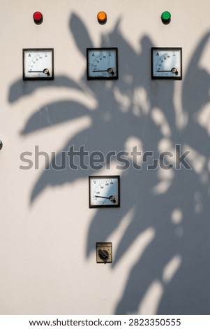 Industrial electrical control panel with analog meters and indicator lights under shaded foliage, used for monitoring and managing power systems.