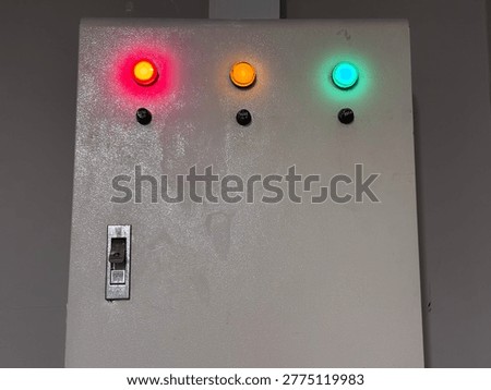 Industrial Electrical Control Panel with Glowing Red, Yellow, and Green Indicator Lights A close-up shot of a metal industrial electrical distribution box featuring three glowing status indicator lights (red, orange, and green) and a main circuit breaker switch. This image represents power management, electrical safety, and industrial automation status