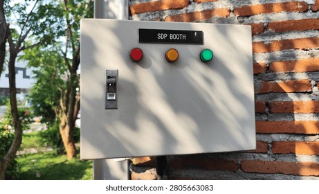 Industrial electrical control panel mounted on a brick wall outdoors. The control box features red, yellow and green indicator lights with a circuit breaker switch, representing electrical systems, industrial automation, power distribution, safety monitoring, and engineering maintenance concepts.