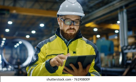 Industrial Engineer In Hard Hat Wearing Safety Jacket Uses Touchscreen Tablet Computer. He Works At The Heavy Industry Manufacturing Factory.