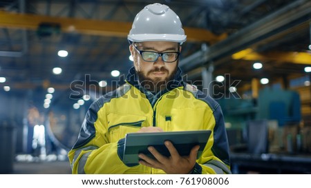 Industrial Engineer in Hard Hat Wearing Safety Jacket Uses Touchscreen Tablet Computer. He Works at the Heavy Industry Manufacturing Factory.