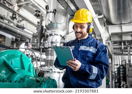 Industrial engineer standing by gas generator in power plant and controlling electricity production on his tablet.