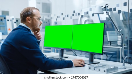 Industrial Engineer Working On A Personal Computer, Two Monitor Screens Are Mock-up, Green Screen, Chroma Key Displays. Modern Factory With High-Tech Machinery