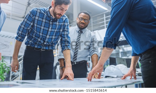 In the Industrial Engineering Facility: Diverse\
Group of Engineers and Technicians on a Meeting Gather Around Table\
Unravel Sheets of Engine Design Technical Drafts, Have Discussion,\
Analyse Drawings