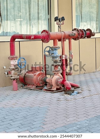 An industrial fire pump system, featuring red-painted pipes and valves installed outdoors on a tiled pavement. the setup includes a large electric motor connected to a pump, with multiple pipes.
