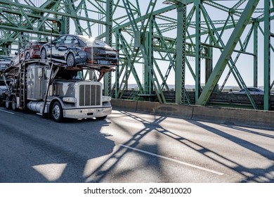 Industrial grade car hauler big rig gray semi truck transporting cars on modular two level hydraulic semi trailer running on the truss arched Interstate Columbia River Drawbridge at sunny day