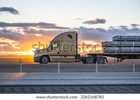 Industrial grade popular big rig beige semi truck tractor with high cab transporting fastened commercial cargo on flat bed semi trailer running on the highway road at sunset in California