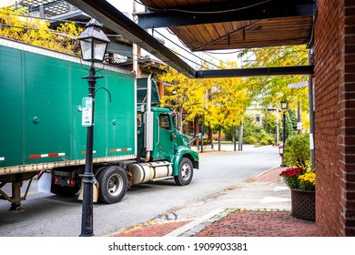 Industrial Green Big Rig Day Cab Semi Truck Tractor With Roof Spoiler Transporting Commercial Cargo In Dry Van Semi Trailer Moving On The Small City Street Road With Buildings In New England