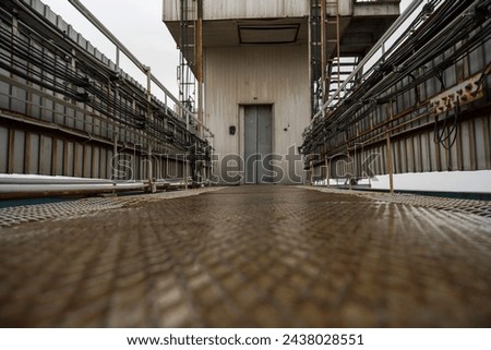 The industrial grit of a metallic catwalk is captured from the ground level, leading the eye to a distant door amidst a complex of pipes and conduits. The perspective emphasizes the scale and utility 