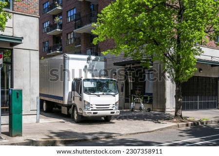 Industrial light duty transportation cab over powerful compact white rig semi truck with big box trailer standing on the urban city street making the local delivery for different customers requests