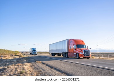 Industrial long hauler big rigs semi trucks team transporting commercial cargo in loaded dry van semi trailers running together on the flat straight highway road in California