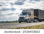 Industrial long hauler carrier beige big rig semi truck with step deck semi trailer transporting fastened plastic pipes running on the summer highway road going between meadows