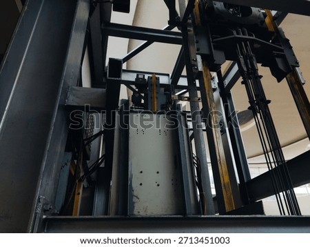 Industrial low-angle view of an elevator counterweight and guide rails inside a shaft, showing steel beams, ropes, and mechanical components.