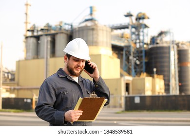 Industrial Male Caucasian Director Talking By VHF Walkie Talkie And Holding Papers Near Factory Outside. Male Wearing White Helmet And Blue Work Jumpsuit. Concept Modern Technology, Plant Profession