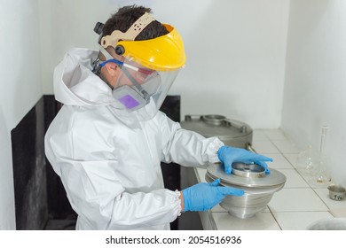 Industrial man worker in white protective overalls goggles, mask and face shield putting the lid on an asphalt test container