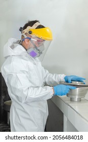 Industrial man worker in white protective overalls goggles, mask and face shield putting the lid on an asphalt test container