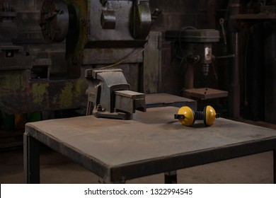 Industrial Mechanical Cast Iron Vice Bolted To A Metal Work Station Table In A Dark Traditional Old Style Repair Workshop Next To Yellow Ear Defenders