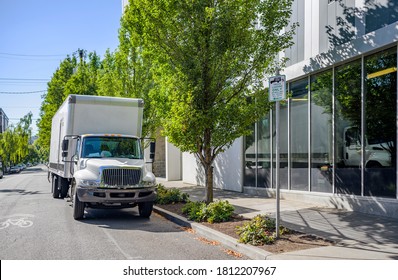 Industrial Medium-sized Professional Big Rig Day Cab Semi Truck With Box Trailer Unloading Delivered Cargo Standing On The Urban City Street With Multilevel Residential Apartment Buildings