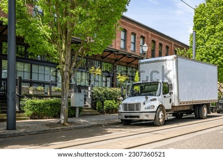 Industrial middle duty day cab powerful compact white rig semi truck with big box trailer standing on the urban city street making the local delivery for different customers needs