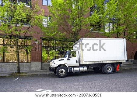 Industrial middle duty day powerful cab compact white rig semi truck with chrome parts and big box trailer standing on the urban city street making the local delivery for different business needs