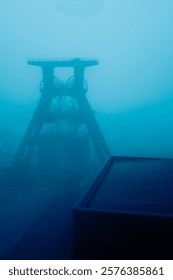 Industrial mining structure emerging through dense blue fog, creating a mysterious and atmospheric composition with sharp lines and minimalistic appeal.